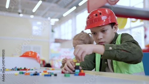 Little African American boy in safe helmet building tower using colorful toy blocks on the table in educational gaming center. Slow motion