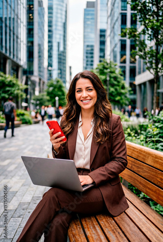 Empresaria sonriente con traje marrón usando un portátil y teléfono en un banco urbano rodeada de edificios corporativos descansando del aire laboral.