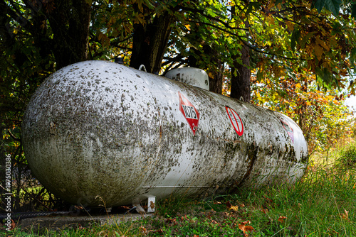 An image of a large unused propane storage tank with warning labels and covered with deal leaves and corrosion. 