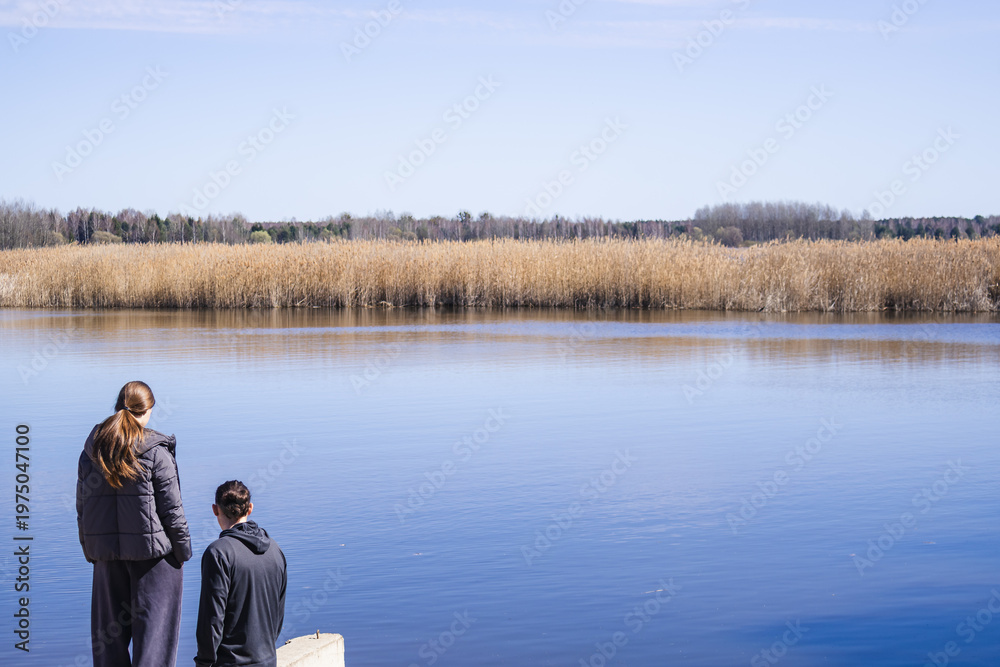 Fototapeta premium Back view of family with father and daughters standing by lake and looking at water together. Warm moment of connection. Concept of family bonding, parenting and peaceful outdoor lifestyle in Poland.