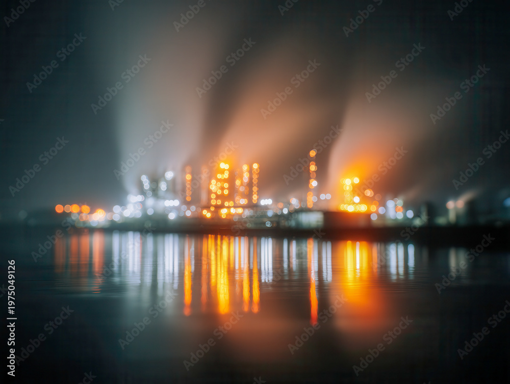 Fototapeta premium Vibrant and glowing industrial plant illuminated at night with bright lights reflecting on calm water surface under a misty sky