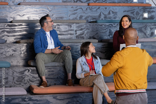 Diverse team sitting on tiered seats discussing using laptop, notes, with mustard jacket, lanyards