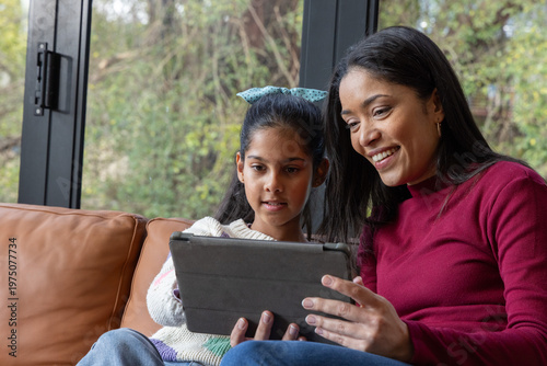 Mother and Indian child sharing tablet on leather sofa beside black-framed window bow visible
