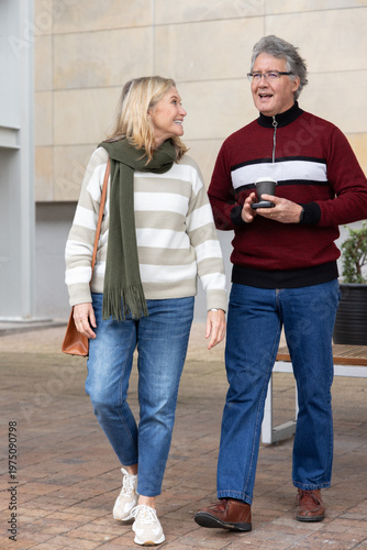Senior couple walking and talking on paved plaza, holding takeaway coffee cup and shoulder bag