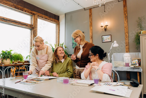 Four women gather in a warm studio filled with plants and natural light. They share laughter and ideas as they work on colorful art projects, enjoying a creative afternoon together.