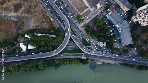 Aerial view of Tbilisi highway interchange traffic flow curving overpasses along riverbank with urban rooftops and vehicles moving across lanes at day time filmed by drone. City scenery from above