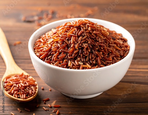 A white bowl filled with reddish-brown grains, and a wooden spoon with rice, sit on a textured, brown surface. Focus is on the bowl