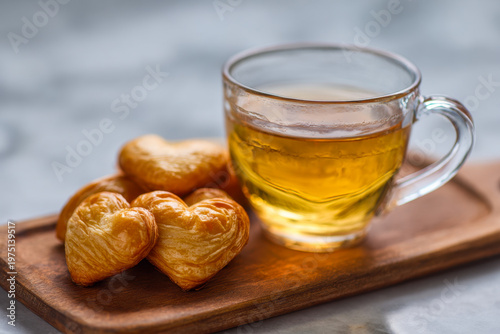 Glass cup of herbal tea served on wooden tray with heart-shaped puff pastries for a cozy and charming afternoon snack or breakfast moment