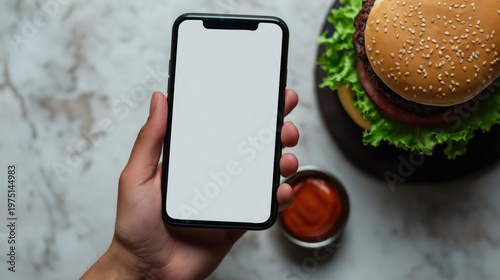 Hand holding smartphone with blank screen near burger and sauce on marble surface