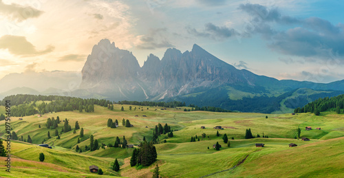 Panoramic view of Alpe di Siusi alpine meadow with green rolling hills, wooden huts and Sassolungo mountains in Dolomites at sunrise, Italy. Rural scenery of Seiser Alm, Dolomiti, South Tyrol
