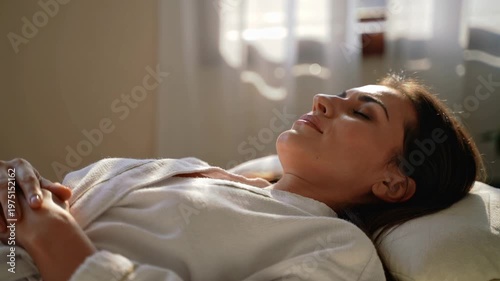 A serene woman lies on a massage table with her eyes closed, hands clasped together, and a peaceful expression on her face.