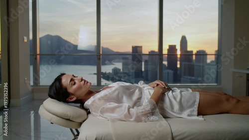 A serene woman lies on a massage table, gazing out the window at a stunning city skyline during sunset, exuding relaxation and tranquility.