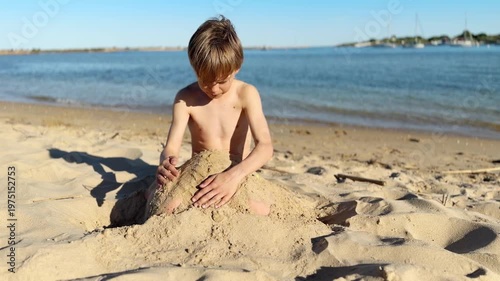 Young boy sitting on warm beach sand carefully shaping a mound near the calm sea under bright sunlight, enjoying summer leisure and creative outdoor playtime by the water