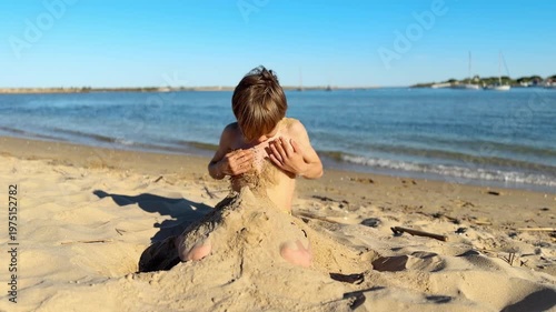 Happy child playing with sand on a sunny beach, enjoying summer vacation by the sea. The barefoot kid is sitting on the shore, building and shaping a sandcastle with his hands