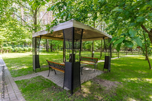 People enjoy sitting on benches under a gazebo in a park. The area is surrounded by green trees. It is sunny and they relax in the afternoon shade