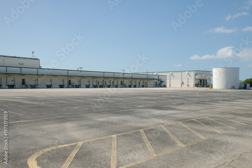 Quadro su tela Large industrial warehouse with multiple loading docks, empty asphalt yard and stair access under a clear blue sky, conveying logistics, transport infrastructure, storage and quiet commercial space