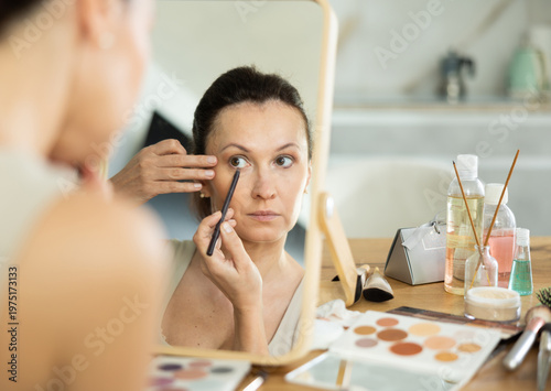 Fotografija Focused adult woman reflected in tabletop mirror, carefully applying eyeliner during daily beauty routine, preparing for day in well-lit room with makeup kit