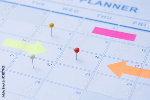 Close up of a monthly wall calendar featuring push pins and bright arrow post-it notes to mark important dates. Concept project management, financial planning, and organized workflow.