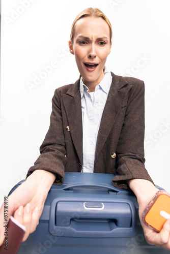 Obraz na plátne Surprised young woman in brown blazer holds blue suitcase and passport, looking down with astonished expression