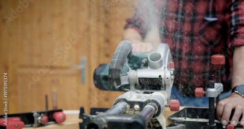 Man uses a miter saw to cut wood for a carpentry project in a workshop during the daytime with dust in the air from the saw