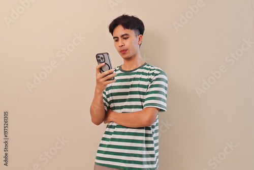 Bild auf Leinwand Asian man using mobile phone with confused expression on plain isolated background, showing uncertainty, digital distraction, or puzzlement while checking smartphone