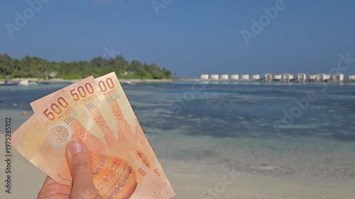 Stack of 500 maldivian ruffiyaa Banknotes in hand with maldivian island and ocean in background. Expencive travel concept