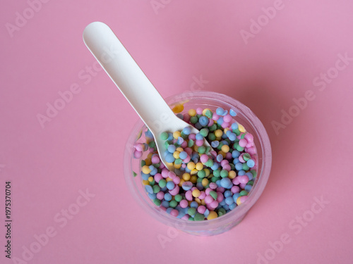 Colorful sugar sprinkles in a small plastic cup with a white spoon on a soft pink background. Minimalist top view composition with vibrant pastel candy balls