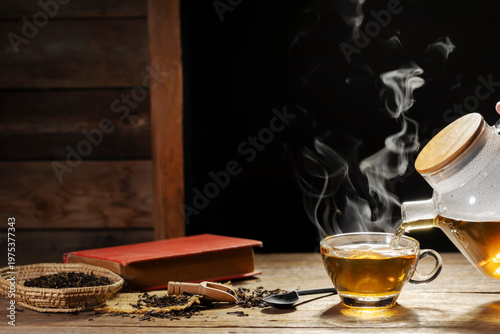 Hot tea pouring into glass cup with book on rustic wooden table.