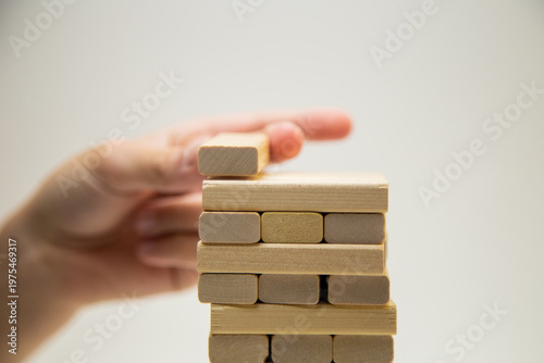 Close-up of the top tower made of wooden bars, a game of jengo, standing on a white background, hand on top placing a wooden block. The concept of board games, business, construction. High quality