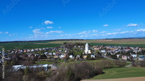 Aerial drone view of Hněvošice in the Czech Republic during a sunny spring day. The image shows a peaceful village surrounded by green fields under a clear blue sky, capturing the beauty of the Czech 
