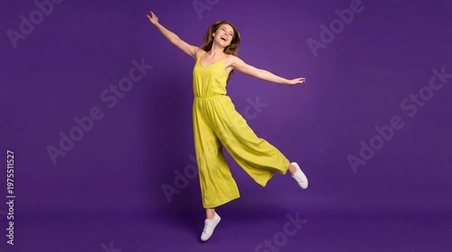 Excited young woman in yellow jumpsuit jumping and dancing over a purple studio background.