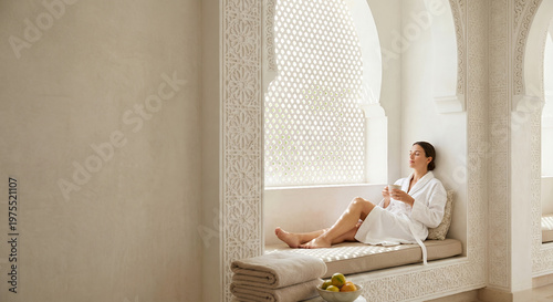 A woman in a white robe sits by a window with her eyes closed, holding a mug and enjoying a moment of relaxation.