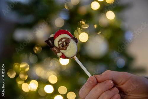 Gros plan d'un enfant ouvrant une sucette en chocolat en forme de tête de père noël, sapin de noël avec guirlande lumineuse en fond.