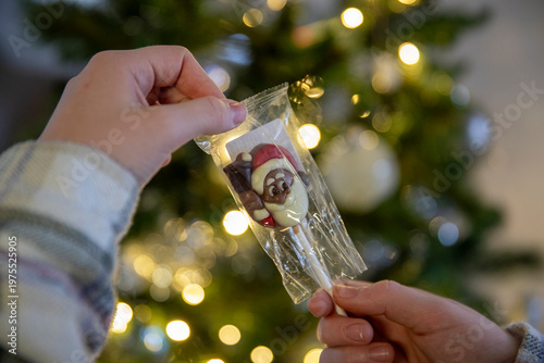 Gros plan d'un enfant ouvrant une sucette en chocolat en forme de tête de père noël, sapin de noël avec guirlande lumineuse en fond.