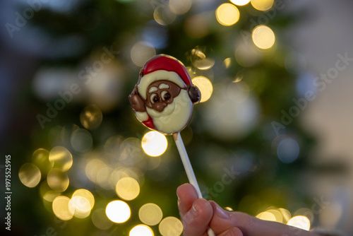 Gros plan d'un enfant ouvrant une sucette en chocolat en forme de tête de père noël, sapin de noël avec guirlande lumineuse en fond.