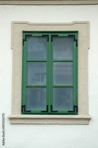 Green wooden window with decorative wrought Iron reinforcement brackets and ecru frame on white wall