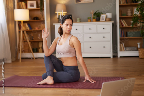 A woman performs a seated twist on a yoga mat while following an online workout on her laptop. This home exercise emphasizes flexibility and core strength in a warm, inviting room.