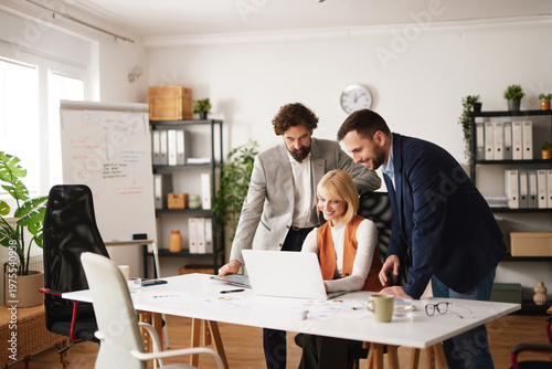 A group of three team members collaborates in an office setting. They gather around a laptop discussing ideas. The atmosphere shows engagement and focus on teamwork.