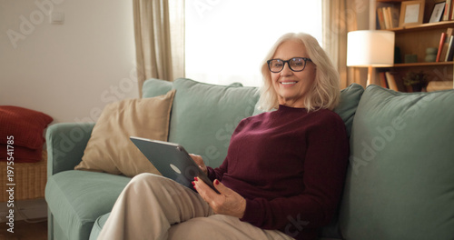 Woman sits comfortably on a couch while using a tablet. Sunlight shines into the room, creating a warm atmosphere as she enjoys her time with the device.