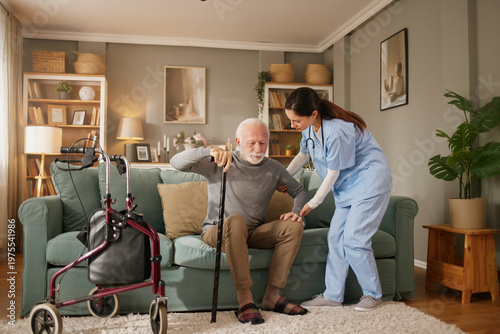 A health worker helps an older man as he stands from a couch in a living room. The space has plants, a walker, and shelves. Both show focus on the activity of support and care.