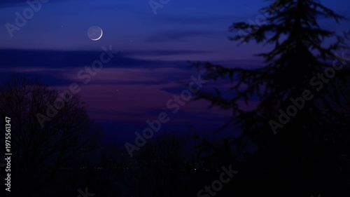 Blue hour time with crescent Moon, stars and planets above landscape silhouettes.