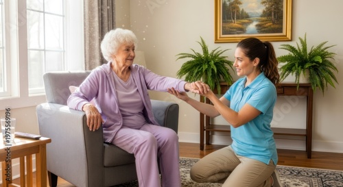 An elderly woman sits in an armchair, holding her hand out to a young caregiver who is kneeling beside her, gently taking her hand. They are in a warmly lit living room with a window 