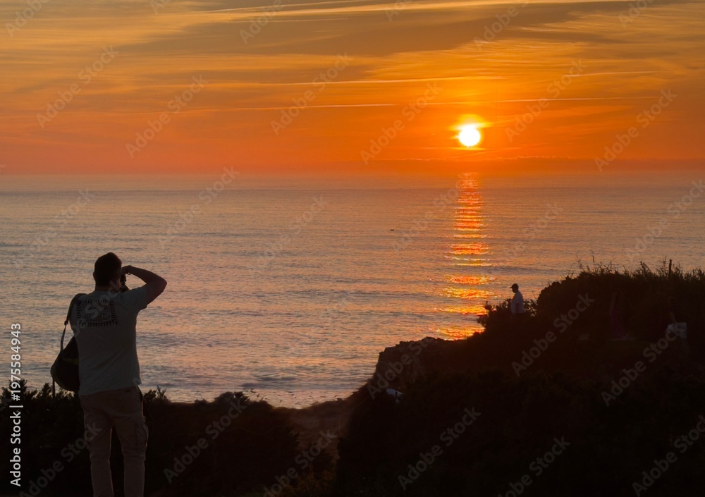 Fototapeta premium Photographer silhouetted on a cliff documenting a vibrant golden hour sunset over the sea