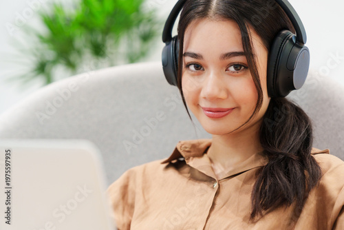 Close up of young, beautiful, happy Asian woman sitting on sofa in cozy, modern living room, listening to music with headphones, smiling, looking at camera with computer laptop