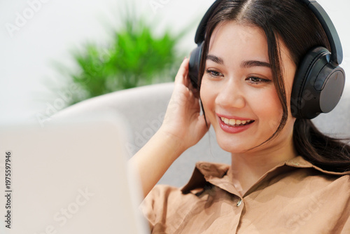 Close up of young, beautiful, happy Asian woman sitting on sofa, listening to music with headphones, smiling, looking at computer laptop at home. Music therapy, lifestyle, technology.