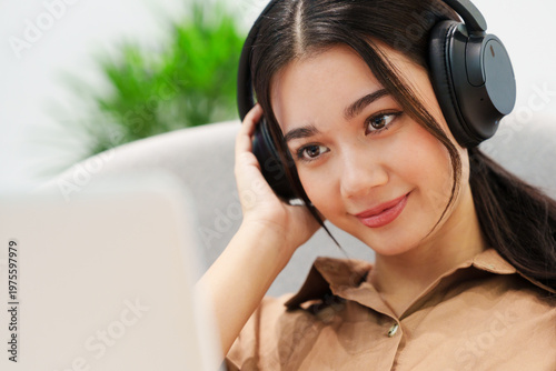 Close up of young, beautiful, happy Asian woman sitting on sofa, listening to music with headphones, smiling, looking at computer laptop at home. Music therapy, lifestyle, technology.