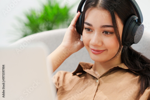 Close up of young, beautiful, happy Asian woman sitting on sofa, listening to music with headphones, smiling, looking at computer laptop at home. Music therapy, lifestyle, technology.