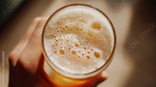 Close up top view of a person's hand holding a golden beer with frothy white foam and bubbles in a clear glass with soft natural light casting shadows on a blurred background