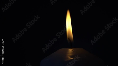 Single candle flame glows softly against pitch black background. Candle flame flickers gently, casting warm, golden glow. Stillness surrounds the flame, evoking quiet peace or solemn reflection