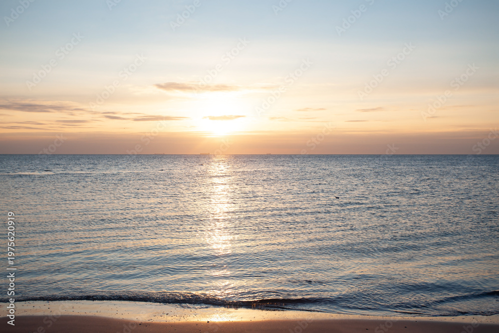 Obraz premium Beautiful landscape of a tranquil sea at sunrise. Soft pastel colors of the sky reflect on the water surface with gentle ripples on the sand. Serene morning atmosphere at the coastline.